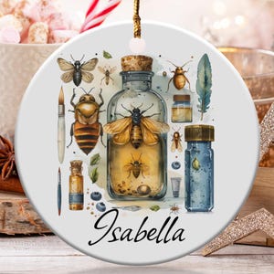 May include: A white ceramic ornament featuring a watercolor illustration of various insects, vials, and a paintbrush. The ornament includes the name "Isabella" in cursive script. The design incorporates shades of brown, yellow, and blue.