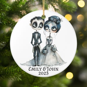 May include: A white ceramic Christmas ornament featuring a skeletal bride and groom in formal attire. The bride wears a white gown and the groom a black tuxedo. The ornament has the text "EMILY & JOHN 2025" in a decorative font. A gold string is attached for hanging.