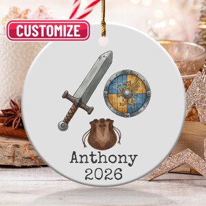 May include: A white ceramic ornament featuring a sword, shield, and pouch design. The shield is blue and yellow with a gold griffin. The ornament has the name "Anthony" and the year "2026" printed on it. A gold string is attached for hanging.