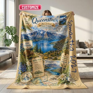 May include: A beige throw blanket featuring a Queenstown, New Zealand travel theme with a mountain and lake landscape, a kiwi bird, and vintage-style postcards. The text includes "Queenstown," "Roys Peak," and the name "Samantha 2027" in blue.