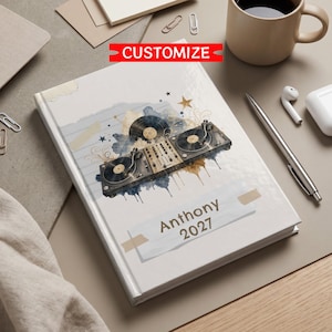 Puede incluir: Un cuaderno de tapa dura blanco con un gráfico de un tocadiscos de DJ y discos de vinilo con detalles en acuarela azul y dorada. La portada incluye el texto "Anthony 2027" impreso en una fuente oscura y una banda roja "CUSTOMIZE" arriba.