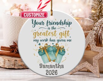 May include: A round white ceramic Christmas ornament with a gold hanging string. The text reads "Your friendship is the greatest gift my work has given me." Two teal gift boxes with gold bows are below the text. The name "Samantha" and the year "2026" are also printed.
