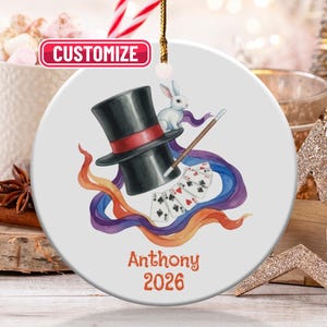 May include: A round ceramic ornament featuring a magician's hat, a white rabbit, playing cards, and a magic wand. The ornament has a black hat with a red band, and the name "Anthony" and the year "2026" are printed in orange.