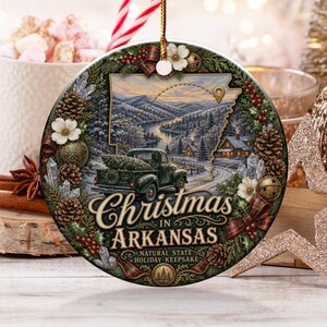 May include: A round ceramic Christmas ornament featuring a green vintage truck carrying a pine tree, set against a snowy Arkansas landscape. The text reads Christmas in ARKANSAS, Natural State Holiday Keepsake, framed by pinecones and berries.
