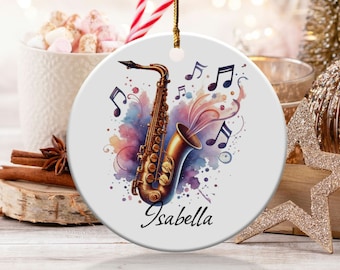 May include: A round, white Christmas ornament featuring a watercolor illustration of a saxophone with musical notes. The saxophone is gold-toned, with colorful splashes of purple, pink, and blue. The name "Isabella" is written in black script.