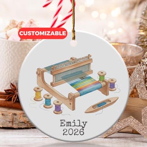 Op de afbeelding: Ronde, witte keramische kerstbal met een aquarel illustratie van een weefgetouw en garenklosjes. De kerstbal heeft het woord "CUSTOMIZABLE" en de naam "Emily" met het jaartal "2026" erop.