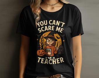 May include: Black t-shirt with a Halloween graphic. The design shows a cartoon witch holding a book and wand, with a pumpkin, tombstone, and the text "YOU CAN'T SCARE ME I'M A TEACHER".