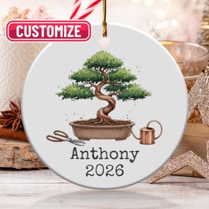 May include: A white ceramic Christmas ornament with a bonsai tree design in a brown pot, with a watering can and scissors. The ornament has the name "Anthony" and the year "2026". The word "Customize" is in a red box at the top.