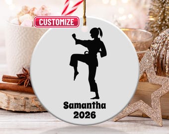 May include: A white ceramic ornament featuring a black silhouette of a person in a karate stance. The name "Samantha" and the year "2026" are printed in black below the figure. The ornament has a gold string for hanging.