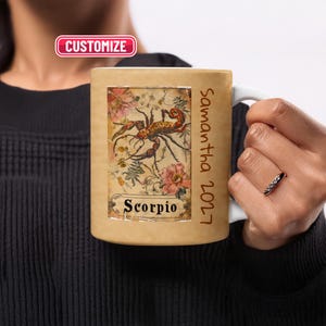 May include: A tan ceramic mug featuring a rectangular graphic of a scorpion surrounded by floral illustrations and the text Scorpio. The side of the mug displays the vertical text Samantha 2027. A pink rectangular label with the text CUSTOMIZE is visible.