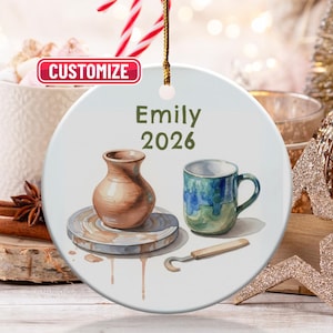 May include: A ceramic ornament with a watercolor design featuring a pottery wheel, a brown vase, and a blue and green mug. The ornament has the name "Emily" and the year "2026" printed on it. A gold string is attached for hanging.