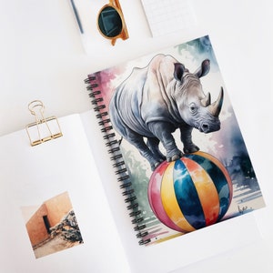 Puede incluir: Una pintura de acuarela de un rinoceronte blanco equilibrándose sobre una bola de colores. El rinoceronte tiene un cuerno grande y está de pie sobre sus patas traseras. El fondo es una suave acuarela de azul y rosa.