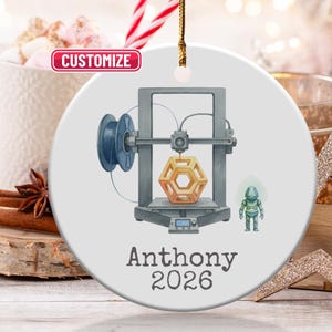 May include: A white ceramic ornament featuring a 3D printer design with a geometric shape. The ornament includes the name "Anthony" and the year "2026". A small green robot is also depicted. The word "Customize" is in a red box.