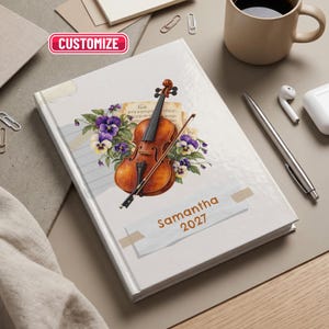 May include: A hardcover notebook featuring a violin illustration, a bow, sheet music, and purple pansy flowers on the cover. The cover includes a white rectangular label with the text Samantha 2027 and a pink button labeled CUSTOMIZE.