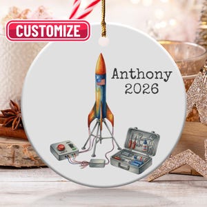 May include: A white ceramic Christmas ornament featuring a colorful rocket ship design with the text "Anthony 2026." The ornament also includes a toolbox and control panel. The word "Customize" is in a red box.