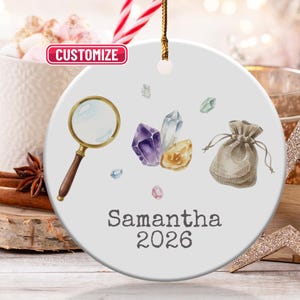 Puede incluir: Adorno de cerámica blanca con una lupa, cristales y una pequeña bolsa. El adorno tiene el nombre "Samantha" y el año "2026" impresos. La palabra "Customize" está en un recuadro rosa.