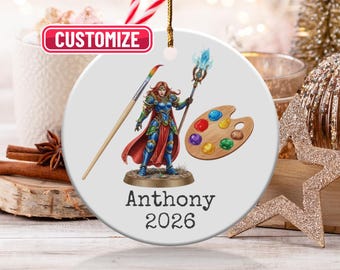 Puede incluir: Adorno navideño de cerámica blanca con un diseño de arte fantástico. El diseño incluye una guerrera con un pincel, una paleta de colores y el nombre "Anthony" con el año "2026". El adorno tiene una cuerda dorada para colgar.