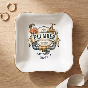 May include: A square white ceramic trinket dish with a gold-toned rim. The center features a graphic of a pipe wrench and faucet with the text PLUMBER, QUALITY WORK BUILT TO LAST, Anthony, and 2027 printed in blue and brown tones.