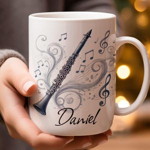 May include: White ceramic mug with a black clarinet design, musical notes, and swirls. The name "Daniel" is written in script. The mug has a white handle. The mug is held by a person.