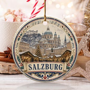 Puede incluir: Un adorno navideño redondo de cerámica con una ilustración detallada del centro de la ciudad de Salzburgo en invierno, incluyendo la catedral y un mercado festivo. El texto SALZBURG aparece en una cinta inferior con notas musicales.