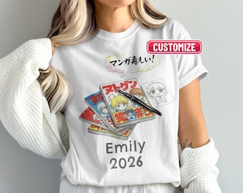 Könnte beinhalten: Weißes T-Shirt mit Manga-Design. Die Grafik zeigt gestapelte Manga-Bücher, einen Stift und eine Skizze eines Manga-Charakters. Japanischer Text befindet sich über den Büchern. Der Name "Emily" und das Jahr "2026" sind darunter gedruckt.