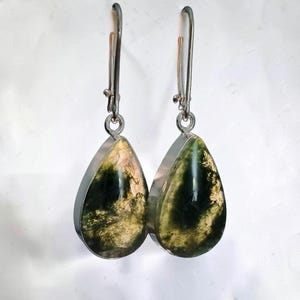 May include: A pair of teardrop-shaped earrings featuring polished green and gold moss-like inclusions encased in a smooth, clear stone setting. The stones are held by silver-toned metal bezels and attached to long, slender silver-toned ear wires.