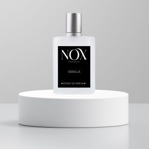 May include: A clear glass bottle of vanilla perfume with a black label that says "NOX FRAGRANCES" and "VANILLA" in white text. The label also says "EXTRAIT DE PARFUM" in smaller white text. The bottle is sitting on a white pedestal.
