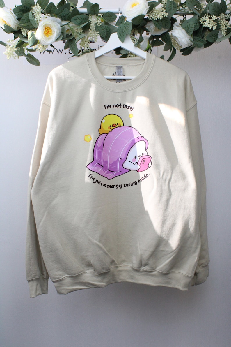 Pu&ograve; includere: Una felpa beige con una grafica di un personaggio dei cartoni animati in una coperta rosa con un pulcino giallo in cima. Il testo sulla felpa recita "Non sono pigro, sono solo in modalit&agrave; risparmio energetico."