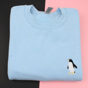 Puede incluir: Sudadera azul claro con cuello redondo y mangas largas. La sudadera presenta un ping&uuml;ino bordado en la parte inferior derecha. El ping&uuml;ino es negro y blanco con pico y patas amarillas. La sudadera est&aacute; doblada.