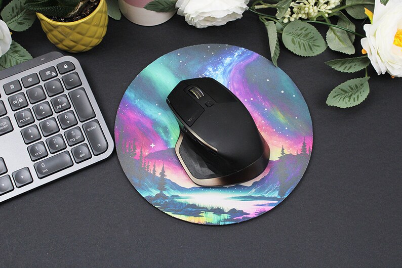 Pu&ograve; includere: Un mouse wireless nero con un accento dorato si trova su un tappetino per mouse rotondo con un design colorato di aurora boreale. Il tappetino per mouse presenta un lago blu scuro con una foresta sullo sfondo.