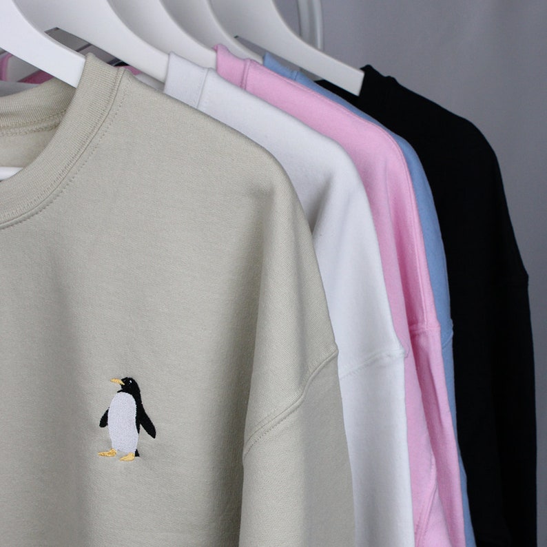 Puede incluir: Una fila de sudaderas de cuello redondo en varios colores, incluyendo beige, blanco, rosa, azul claro y negro, colgadas en perchas blancas. La sudadera beige presenta un dise&ntilde;o de ping&uuml;ino bordado.