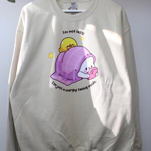 Pu&ograve; includere: Una felpa beige con una grafica di un personaggio bianco avvolto in una coperta viola con un pulcino giallo in cima. Il testo sulla felpa recita "Non sono pigro, sono solo in modalit&agrave; risparmio energetico."