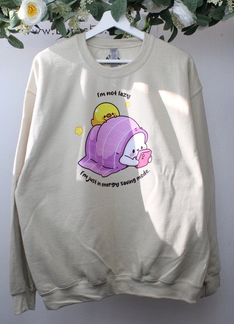 Pu&ograve; includere: Una felpa beige con una grafica di un personaggio bianco avvolto in una coperta viola con un pulcino giallo in cima. Il testo sulla felpa recita "Non sono pigro, sono solo in modalit&agrave; risparmio energetico."