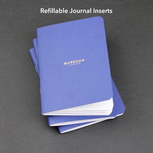 Könnte beinhalten: Ein Stapel blauer, nachfüllbarer Journal-Einsätze mit weißen Seiten. Der Einband des oberen Journals trägt den goldenen Text "BURECHO LONDON". Der Text "Refillable Journal Inserts" befindet sich oben im Bild.