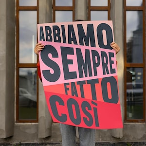 Könnte beinhalten: Eine Person hält ein großes Plakat mit dem Text "ABBIAMO SEMPRE FATTO COSI" in schwarzen Buchstaben auf einem rosa, roten und orangenen Hintergrund.