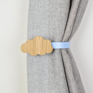 Könnte beinhalten: Ein Vorhanghalter mit einem hellbraunen, wolkenförmigen Holzornament, das an einem glatten, einfarbigen hellblauen Band befestigt ist. Der Halter umschließt einen grauen Stoffvorhang mit einem sichtbaren Fischgrätenmuster und Textur.