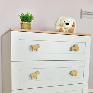 May include: A white dresser with wooden animal-shaped drawer pulls. The dresser has a light wood top and a plush dog toy on top. A small plant in a woven basket sits on the dresser.