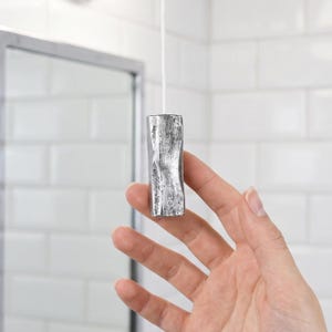 Op de afbeelding: Een zilverkleurige, cilindrische trekschakelaar met een getextureerde, ruwe oppervlakteafwerking. Het object is bevestigd aan een dun wit koord dat verticaal omhoog loopt, met een licht concaaf midden en een metalen, verweerd uiterlijk.