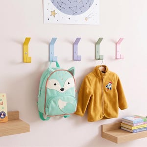 May include: Five wall-mounted metal hooks in yellow, light blue, lavender, sage green, and pink. A mint green fox-shaped backpack hangs from the blue hook, and a mustard yellow zip-up fleece jacket hangs from the green hook on the wall.