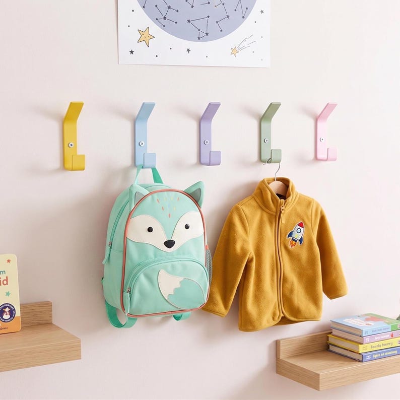 May include: Five wall-mounted metal hooks in yellow, light blue, lavender, sage green, and pink. A mint green fox-shaped backpack hangs from the blue hook, and a mustard yellow zip-up fleece jacket hangs from the green hook on the wall.