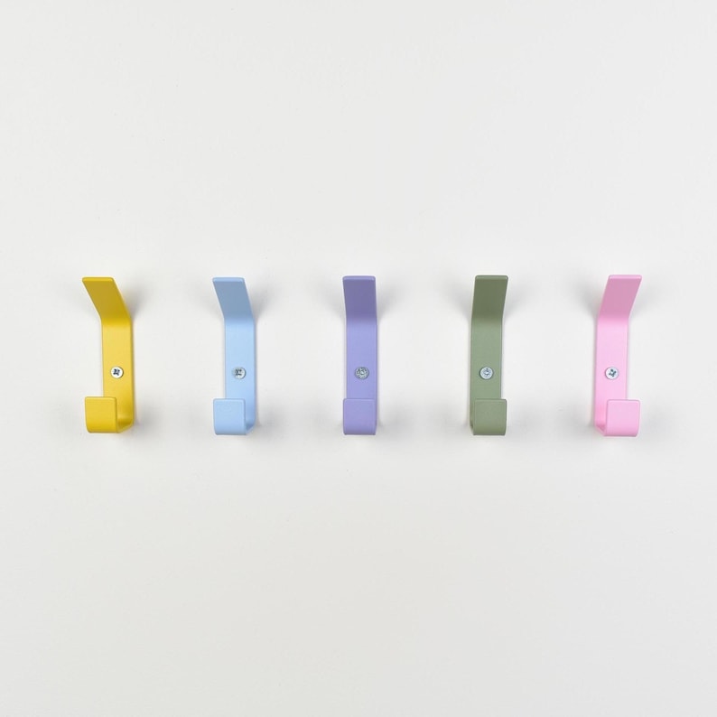 May include: A set of five wall-mounted metal hooks arranged in a horizontal row. Each hook features a distinct matte color: yellow, light blue, lavender, sage green, and pink. Every hook is secured to the wall with a single visible silver screw.