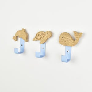 Könnte beinhalten: Drei Wandhaken mit hellen Holzfiguren in Form eines Delfins, einer Schildkröte und eines Wals. Jede Holzfigur ist an einem hellblauen Metallhaken mit einer sichtbaren silbernen Schraube befestigt und bietet eine funktionale Aufbewahrung.