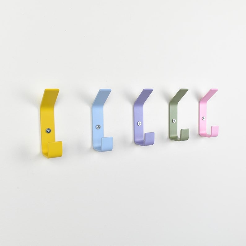 May include: A set of five wall-mounted metal hooks in a row, featuring a matte finish in yellow, light blue, lavender, sage green, and soft pink. Each hook has a simple angled design and is secured to the wall with a single visible silver screw.