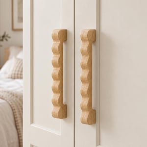 Può includere: Due maniglie verticali per armadi realizzate in legno chiaro con un profilo ondulato e smerlato. Le maniglie sono montate su una coppia di ante di armadio bianche opache e lisce, mostrando una venatura del legno naturale.