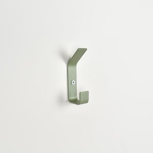May include: A sage green metal wall hook with a dual-prong design, featuring a flat, angled upper arm and a shorter, upright lower hook. The piece is secured to the wall with a single visible silver-toned screw centered on the vertical base plate.