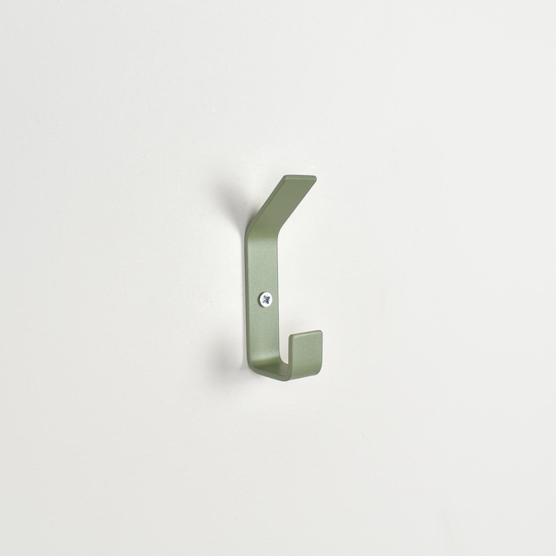 May include: A sage green metal wall hook with a dual-prong design, featuring a flat, angled upper arm and a shorter, upright lower hook. The piece is secured to the wall with a single visible silver-toned screw centered on the vertical base plate.