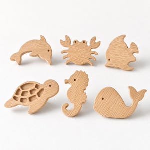 Könnte beinhalten: Ein Set aus sechs hellen Holz-Möbelknöpfen in Form von Meerestieren, darunter ein Delfin, eine Krabbe, ein Fisch, eine Meeresschildkröte, ein Seepferdchen und ein Wal. Jedes Stück weist eine natürliche Holzmaserung und ein kleines Loch auf.