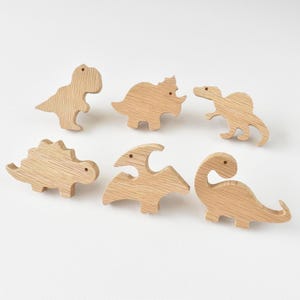 Op de afbeelding: Een set van zes lichtgekleurde houten dinosaurusvormige knoppen, elk met een eigen silhouet waaronder een T-Rex, Triceratops, Spinosaurus, Stegosaurus, Pterodactylus en Brachiosaurus, allemaal met een zichtbare natuurlijke houtnerfstructuur.