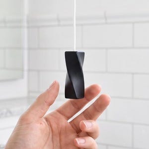 May include: A matte black light pull with a twisted, hourglass-inspired geometric shape. The solid, dark-colored object is attached to a thin white cord, featuring a smooth, uniform finish and a distinct spiraling design that tapers toward the center.