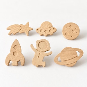 May include: A set of six light-colored wooden wall hooks or decorative pieces featuring space-themed shapes including a shooting star, a flying saucer, a cratered moon, a rocket ship, an astronaut, and a planet with rings, all with a smooth finish.